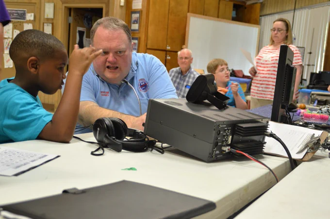 An instructor from NASA assists a young student in using amateur radio equipment during an educational event, with other participants in the background.

📡🎙️ Calling all radio enthusiasts! Join us on June 22 at Highland Road Park Observatory for the American Radio Relay League Field Day! 📻 The Baton Rouge Amateur Radio Club will set up temporary stations and participate in a nationwide emergency exercise. 🌞🔋 Experience the thrill as they use battery and solar power to make contact with other stations across North America! 🚀🌍
	It’s a day of skill, suspense, and fun! 🛰️🌐
