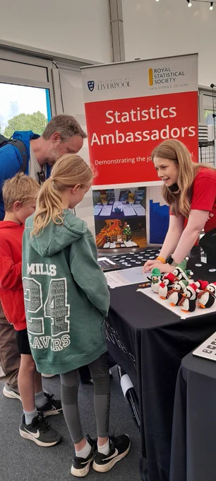 3 children are talking to the science festival host behind a table. On top of the table are Penguins and a maths game. 