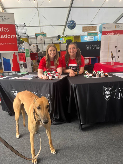 A stall at a science festival. Two tables with a display of Penguins on the table. Two people are smiling at the camera and a dog is infront of the table. 