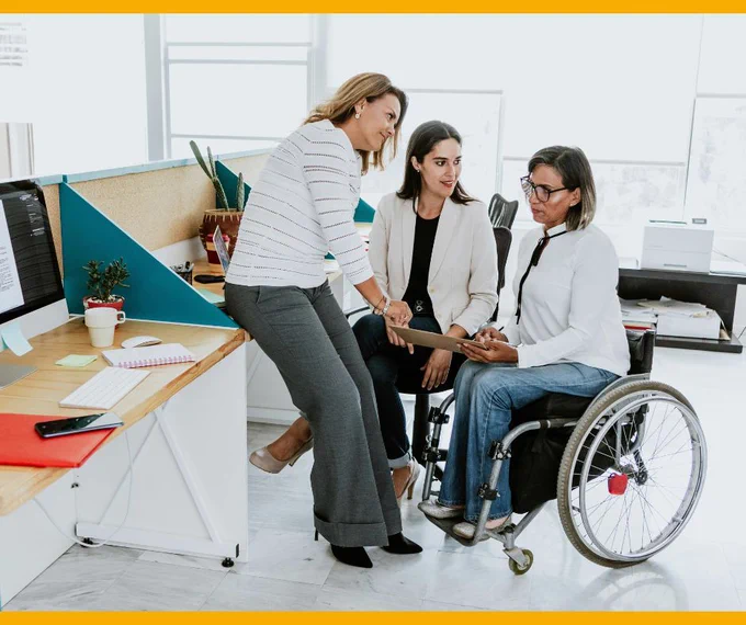 Three people having a conversation in a circle in an office. One person is resting on a desk while another sits in a wheelchair. Another woman is sitting in a chair.