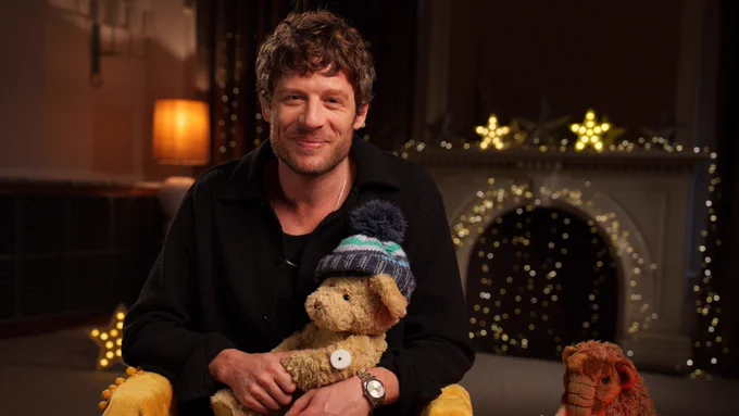 James Norton smiling to camera holding a bear with a glucose monitor on its arm. background of fairy lights and stars around a fireplace
