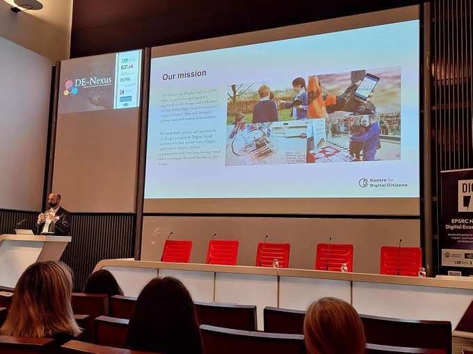 A picture of Dave Kirk giving his talk at the event, he is on the left, and there is a big screen showing images from the project.