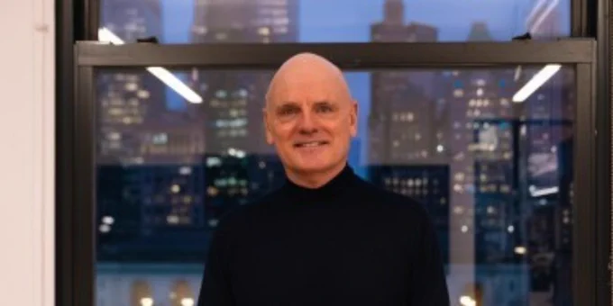 James Corner smiling in front of a window with the New York skyline at dusk.