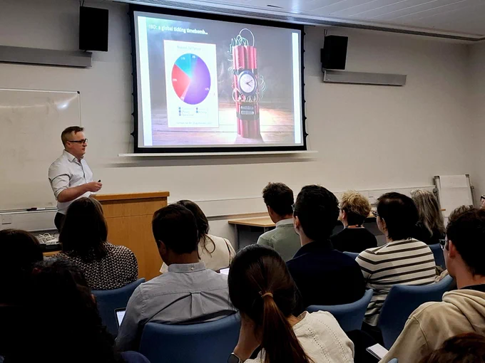 Dr James Lee presents a slide to a group of people in a lecture theatre. The slide reads 'IBD: a global ticking timebomb'.