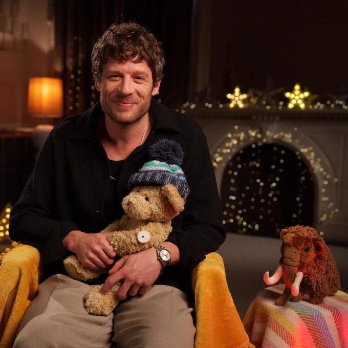 James Norton smiling, seated with a stuffed bear in lap and a plush mammoth toy beside him, in a cosy room with soft lighting and twinkling lights in the background.