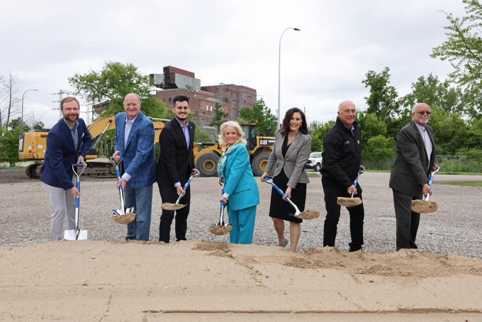 Governor Gretchen Whitmer participates in a groundbreaking for DTE’s new battery facility in Trenton. She is joined by six other people, all of whom are holding shovels. From left to right, they are: Trenton Mayor Steven Rzeppa, DTE Vice Chairman and Group President Trevor Lauer, State Senator Darrin Camilleri, Representative Debbie Dingell, Governor Whitmer, DTE Energy CEO Jerry Norcia, and Wayne County Executive Warren Evans. In the background is various construction equipment used on-site.