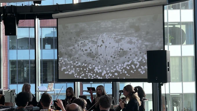 women in a string quartet with a women conductor playing synths. The are playing to a live audience. Behind them on the screen is an animation of a cartoon drawing of a head and lots of black and white dots.