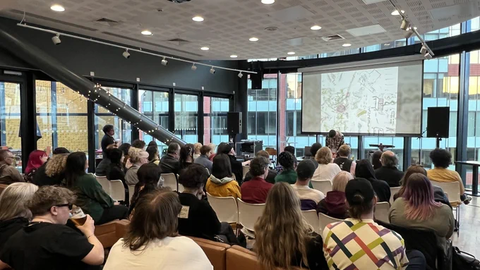 Man playing electronic music to a full audience with a hand drawn image of maps and locations behind him.