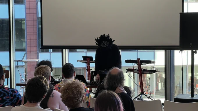Man dressed in an all black costume with a feather mask. Standing in from of a live audience playing electronic music. There are synths on stands behind him.