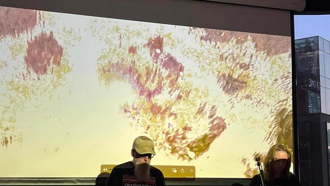 Man with beard in baseball cap and woman in shadow. They are playing electronic instruments. There is a screen with a bright orange and grey filter over running water. 