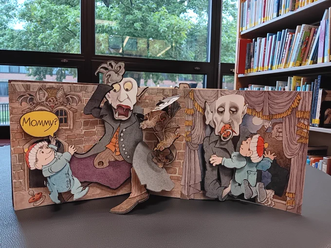 Pop-up book, "Mommy?" open in front of a backdrop of windows and bookshelf. 
