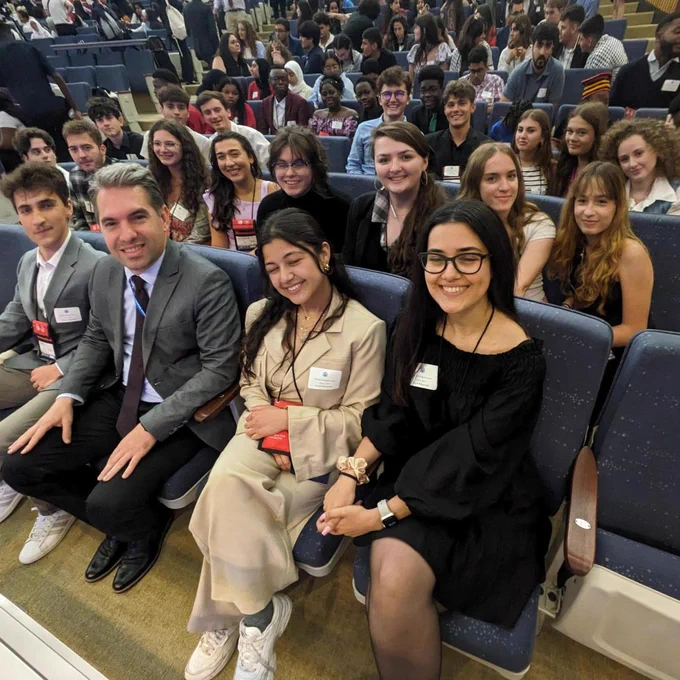 Group of students sitting in an auditorium, smiling and interacting cheerfully, dressed in formal and business casual attire. They are sitting with Diplomat Gent Gjikollori from Kosovo
