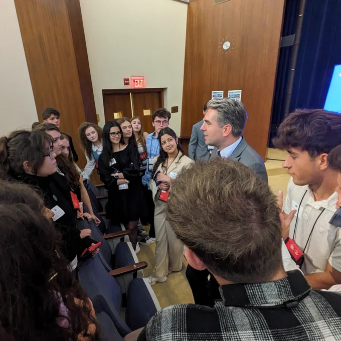 Group of people engaging in a discussion with Diplomat Gent Gjikollori inside a conference room or hall with visible name badges.