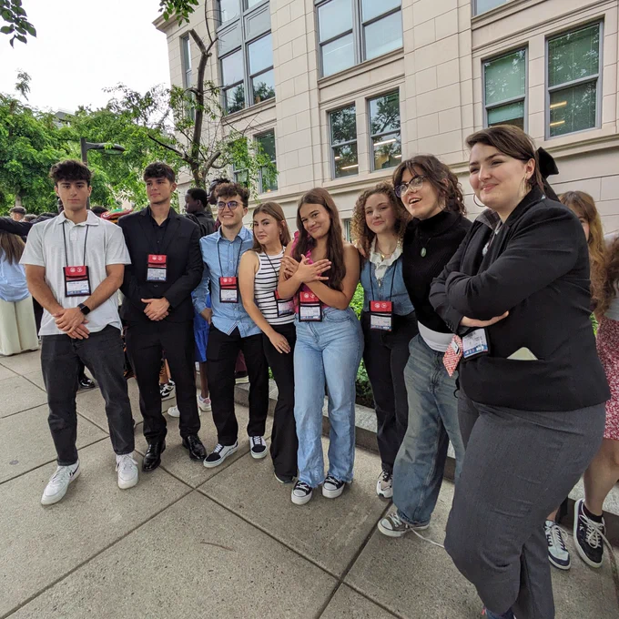 A group of eight young exchange students wearing lanyards, standing together outside a building, smiling and posing for a photo.