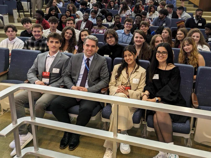 Group of people sitting in a lecture hall, with three individuals in suits in the front row prominently displayed. Everyone appears to be attentively facing forward, posing for photo with Kosovar Diplomat