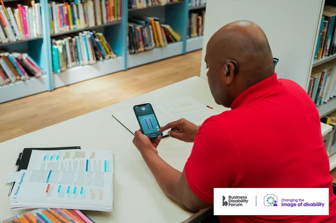 Deaf person wearing a hearing aid using hearing aid software on their phone in a library. The Business Disability Forum 'Changing the image of disability' logo is in the lower right corner of the image.