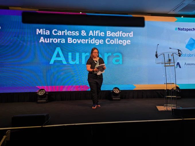 Mia, a short white woman with brown hair and dressed in a black t-shirt, speaks from her notes at the Natspec stage.
