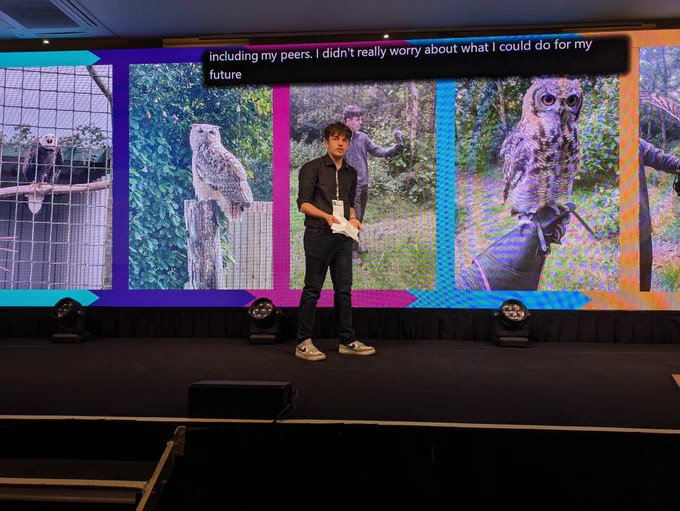 Alfie, a young white man with dark hair, dressed in a black shirt, speaks from his notes on the Natspec stage.
