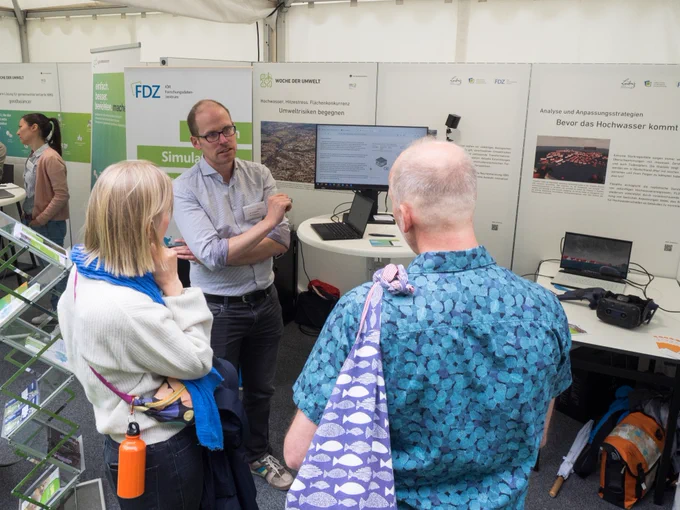 Blick auf den Stand des IÖR auf der Woche der Umwelt. Im Hintergrund Tafeln mit Bildern und Texten zu den Themen des Standes (Hochwasser, Hitzestress, Flächenkonkurrenz). Davor Tische mit Monitoren und einer VR-Anwendung. Im Vordergrund ein Mitarbeiter der IÖR im Gespräch mit Besucher*innen des Standes.