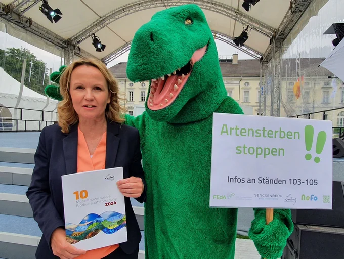 Der Dinosaurier „George“ des Leibniz-Forschungsnetzwerks Biodiversität übergibt die Broschüre mit den „10 Must Knows“ der Biodiversitätsforschung an Bundesumweltministerin Steffi Lemke. 
