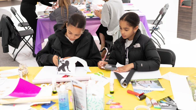 Two individuals focused on crafting at a table covered with colourful papers and art supplies. 