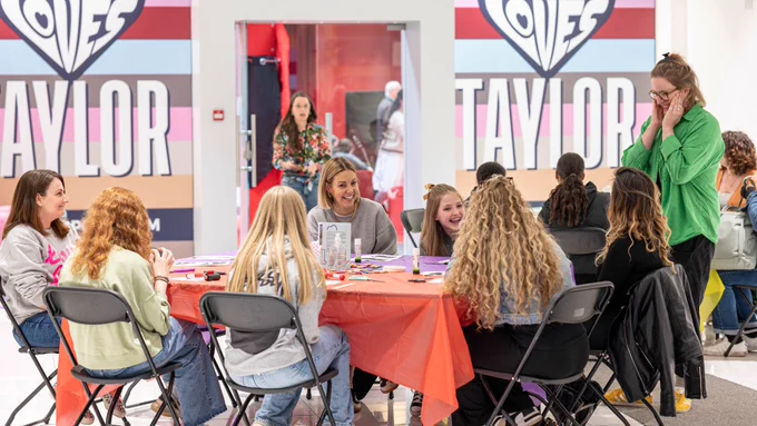A group of people is engaging in a lively activity at a crafting table, surrounded by vibrant "Liverpool Loves Taylor" branded banners, in an indoor public setting.