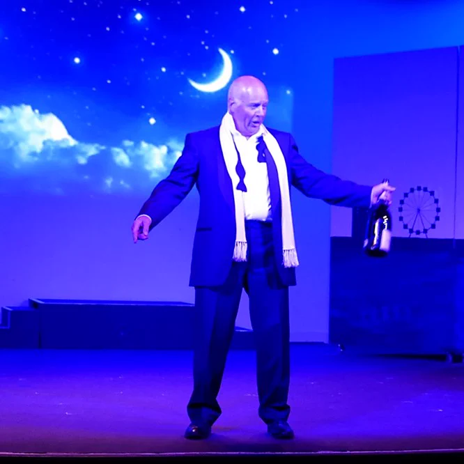 A character from Cheshire Cats. He is dressed in an evening suit, with a loosened black bow tie and a white silk scarf. He is holding a champagne bottle.