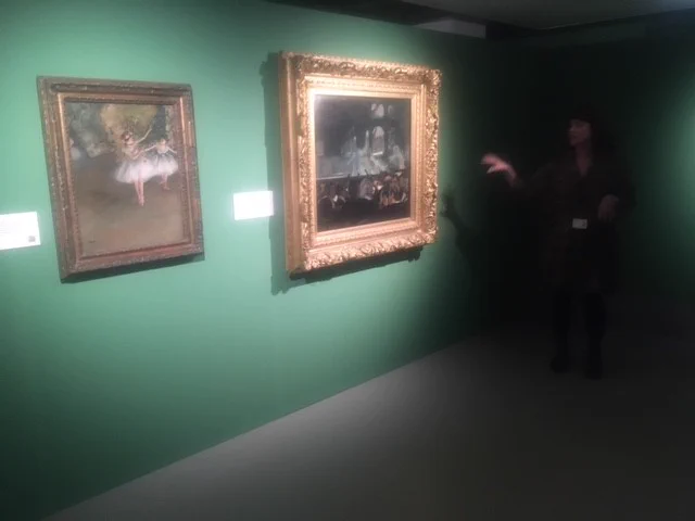 Two framed pictures by artist Edgar Degas hanging on a green wall with a woman to the right of them talking and gesturing. 