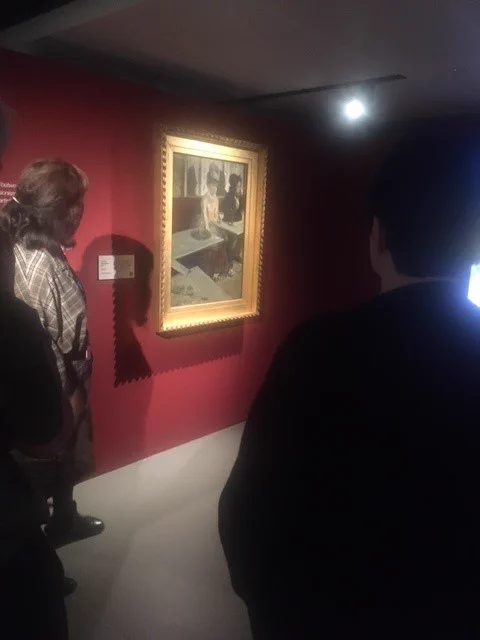Two people, seen from behind, regarding the artwork "L'absinthe" by Edgar Degas hanging on a red wall. 