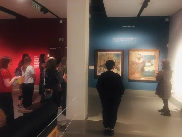 A group of people seen from behind regarding two art work by Edgar Degas, hanging on a blue wall. 