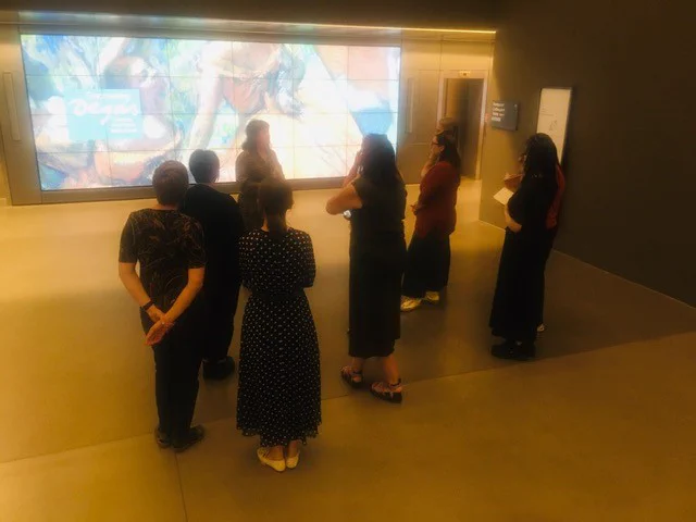 A group pf people seen from behind; listening to a woman talking to them. In front of them a large digital display showing dancers drawn by Edgar Degas. 