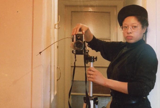 Colour image of a Black woman taking a self portrait in a mirror in the hallway of a flat. She is holding a camera steady on a tripod, wearing a black turtleneck, black beret and glasses.