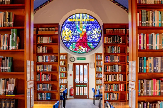 Bookshelves in a light library, with a stained glass window and gilt ceiling.