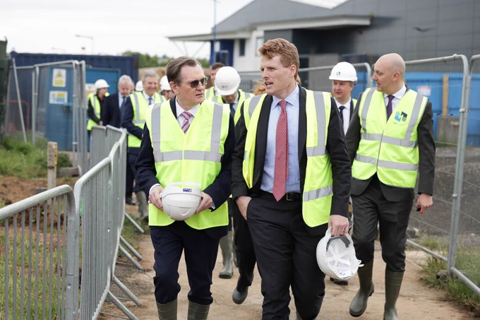 Image shows US Envoy Joe Kennedy and Professor Ian Greer walking across the site wearing high-vis vests and holding helmets, accompanied by other representatives from industry and government.