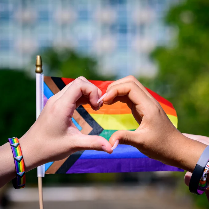 Hands clasped in front of a pride flag against a blurry background of trees. 