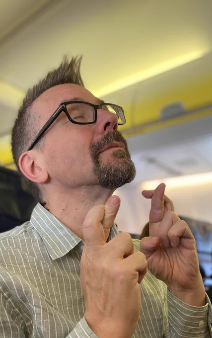 Photo of Alex on a plane with his eyes closed looking upwards with both index fingers crossed