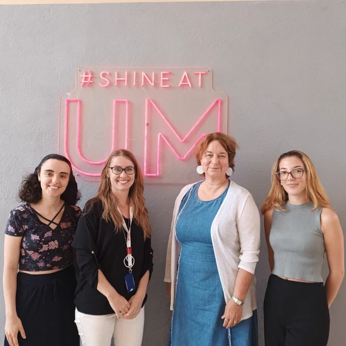 Picture showing 4 women smiling at the camera. A neon sign hanging from the ceiling says #Shine at UM