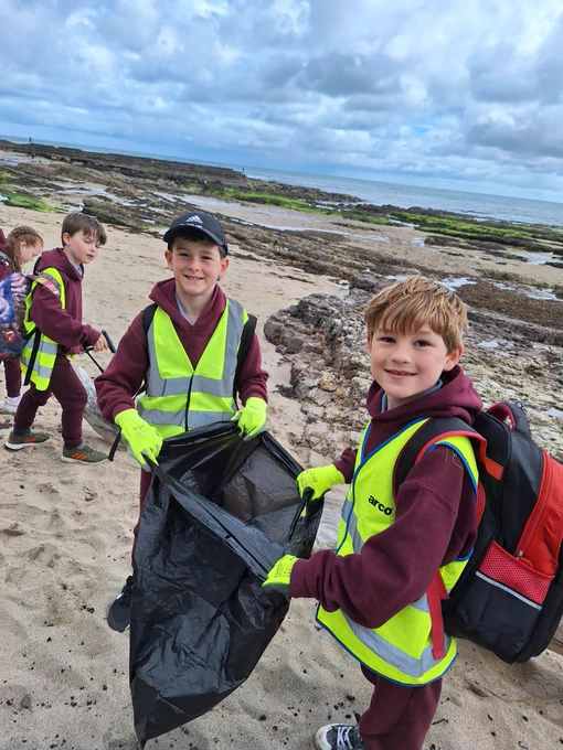 Two smiling children are holding a rubbish bag on a beach, behind them are two children with litter pickers looking for rubbish.