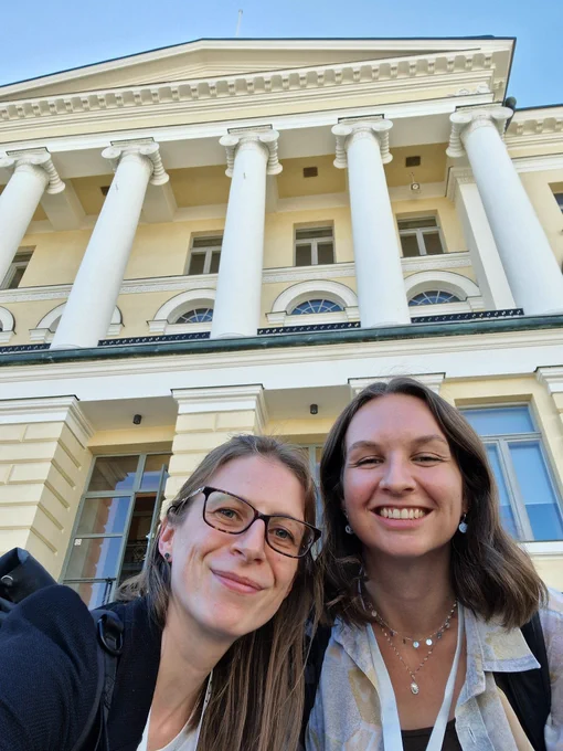 The faces of two women in front of a pale yellow and white neoclassical building with blue sky above 