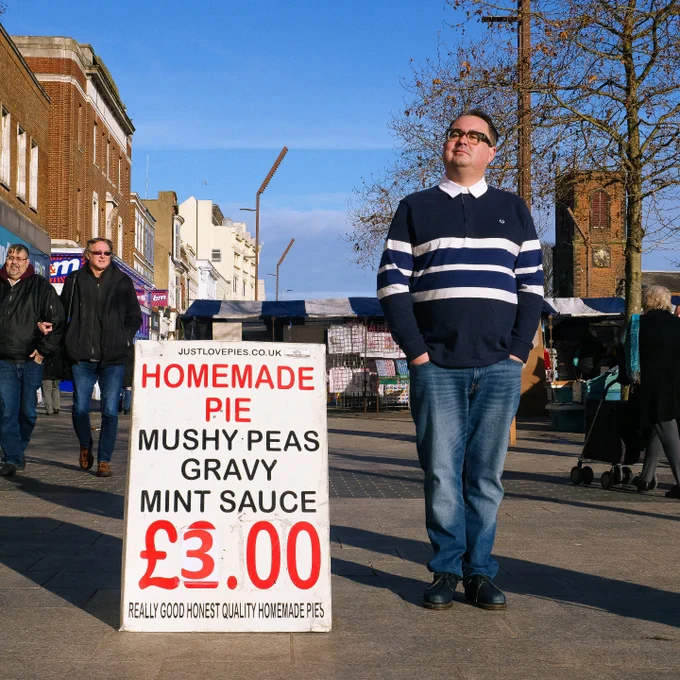 Tommy (he/they) is standing next to a sign on the pavement. Tommy is wearing brown square glasses, blue jeans and a dark blue collared jumper with three big white stripes across the middle, and is looking thoughtfully upwards and towards the right. With black and red lettering on a white background, the sign advertises homemade pies with mushy peas, gravy and mint sauce for £3. A few people can be seen bustling among the shops behind the sign. The natural lighting is warm, clear and bright.