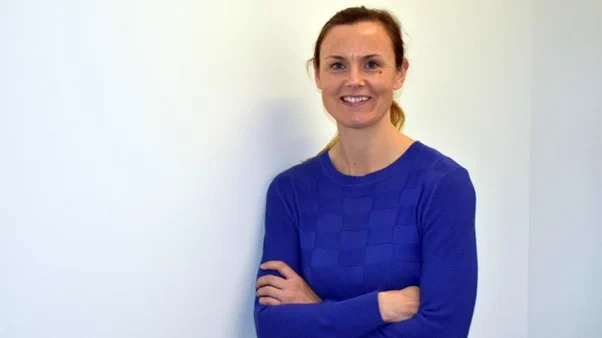 Scholar and former England Cricketer Laura MacLeod stands smiling at the camera with crossed arms, wearing a blue sweater against a plain white wall background.