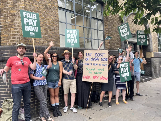 Smiling journalists on picket line wave and hold placards with text fair pay now and 'UK cost of living crisis. Here's how to help, scientist reveal the solution, fair pay now.'