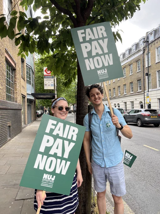 Journalists smile as they hold green placards with white text Fair pay now, NUJ logo in white beneath text. One person wears sunglasses.