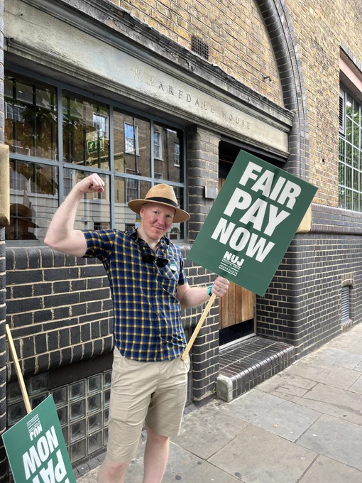 Journalist wearing checked shirt and beige shorts flexes bicep whilst holding green placard with white text 'fair pay now'. NUJ logo in white beneath text. 