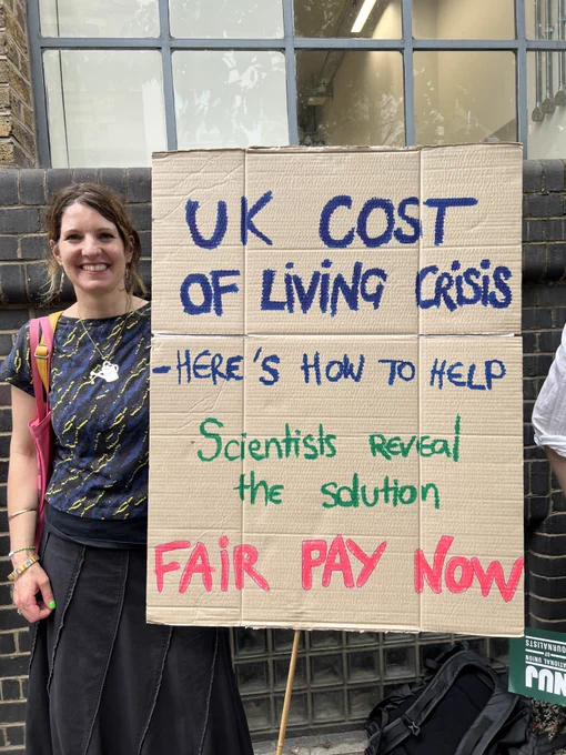 Journalist smiles whilst holding cardboard handmade sign in blue green and pink text 'UK cost of living crisis - here's how to help. scientists reveal the solution, fair pay now.