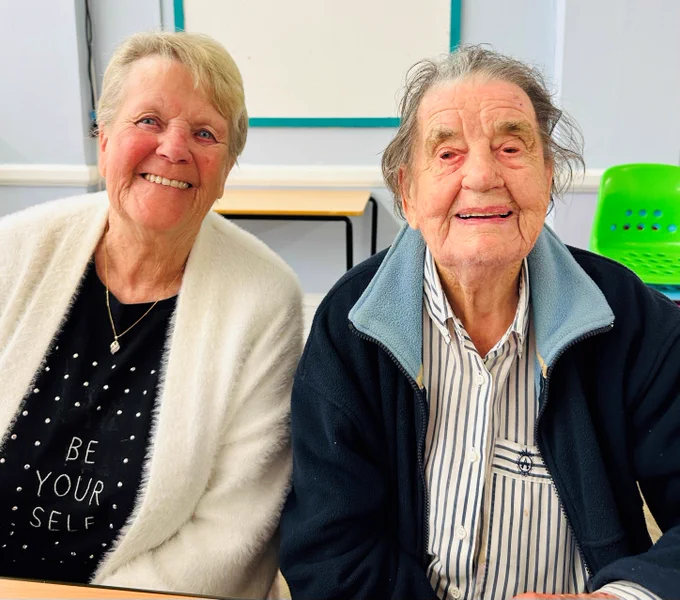 Two ladies smile at the camera, the lady on the left is wearing a white cardigan and has short hair, the lady on the right is wearing a blue jacket and has short brown hair. They are sitting in a room with blue walls.