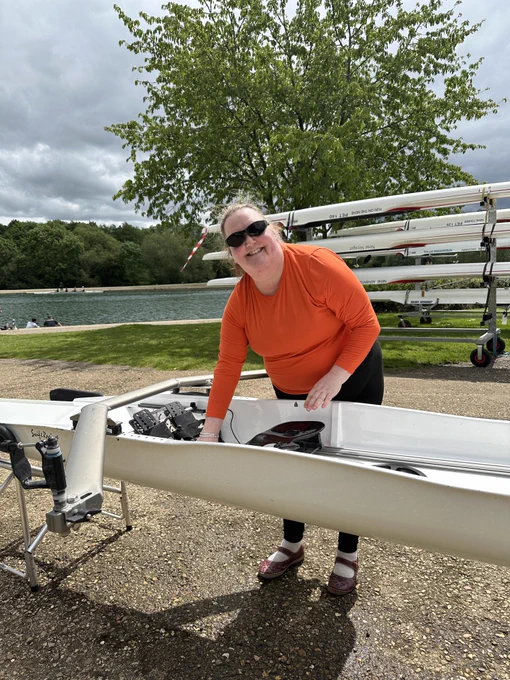 A lady looks at the camera smiling with wearing tinted glasses and a bright orange top. She is standing with her hand cleaning a rowing boat and the sun is shining. Behind her is a rowing lake and boats stacked on to a tailor. 