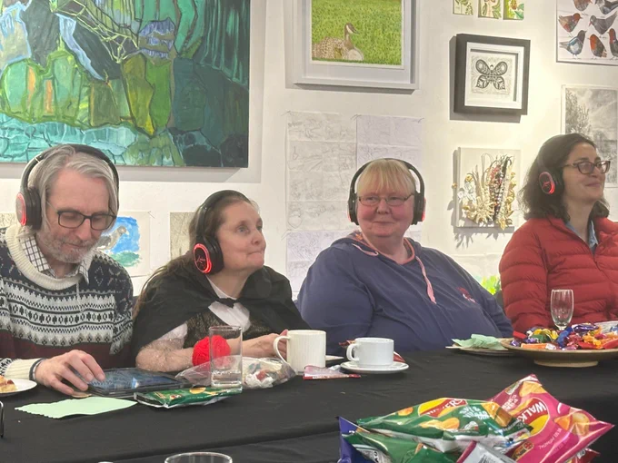 A row of four people sits along a table smiling, they are wearing headsets as they listen to a recording. They are in a museum, so you see large paintings behind them. There are cups of tea and sweets on the tables.
