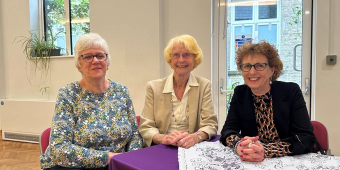 Three ladies sit behind a coffee table in a community hall. They are smiling at the camera, the lady on the right-hand side is Rosie and she wears a black jacket and polka dot dress. 