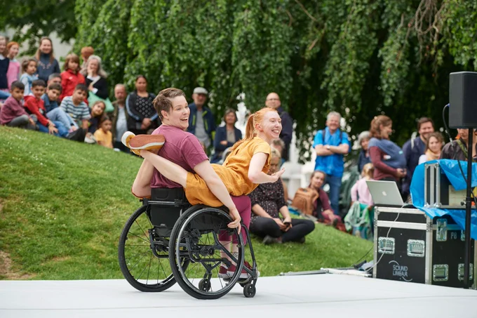 [main image] the ‘Look Mum, No Hands!’ duo performing in front of an audience at Buildstrung festival Germany. The disabled acrobat dressed in burgundy, is balancing the non-disabled acrobat in yellow on their wheelchair. the acrobat in yellow, has their legs wrapped around the disabled acrobat and their chair as they both glide across the stage, smiling at the audience. 